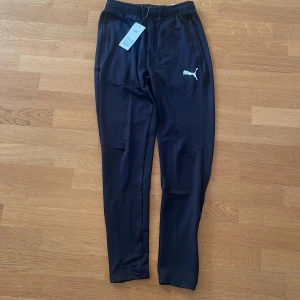 Svarta träningsbyxor från Puma S - Svarta träningsbyxor från Puma med vit logga på låret. Aldrig använda. Slim fit-modell med dragkedjor vid bensluten och elastisk midja med snörning. Tillverkade i syntetmaterial som andas, perfekta för träning eller chill. Snygg och sportig look.