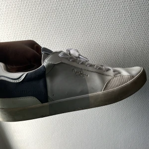 Vita och marinblå sneakers från Pepe Jeans - Snygga sneakers från Pepe Jeans i vitt och marinblått med klassisk design. Skorna har rund tå, vita snören och detaljer i skinn och mocka. Pepe Jeans-logga i silver på sidan och mörkblå detalj på hälen. Perfekta för en stilren och avslappnad look.