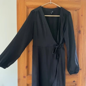 Svart omlottklänning med knyt - Svart omlottklänning från Vero Moda med långa ärmar och knyt i midjan. Klänningen har v-ringning och är tillverkad i ett lätt, mjukt material som faller snyggt.