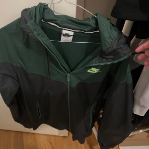 Nike vindjacka i mörkgrönt och svart - Nike vindjacka i mörkgrönt och svart med dragkedja framtill och huva. Jackan har en sportig look med Nike-logga på bröstet och praktiska fickor. Perfekt för dig som gillar streetwear och vill ha en lätt jacka till vardags.