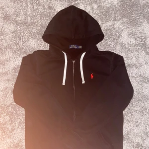 Svart hoodie från Polo Ralph Lauren - Svart hoodie från Polo Ralph Lauren med vit snörning i huvan och dragkedja framtill. Klassisk röd broderad logga på bröstet. Mjuk bomullskänsla och ribbade muddar vid ärmslut och nederkant. Perfekt för chill dagar.