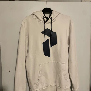 Vit hoodie från Peak Performance M - Säljer en stilren vit hoodie från Peak Performance i storlek M. Tröjan har svart logga framtill, svarta snören i huvan och en klassisk känguruficka. Perfekt för dig som gillar enkel och clean streetstyle.