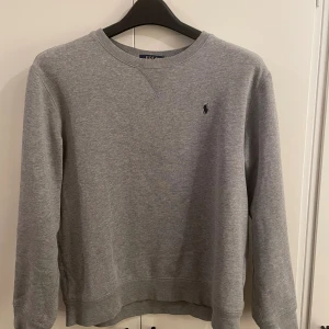 Grå sweatshirt från Polo Ralph Lauren - Klassisk grå sweatshirt från Polo Ralph Lauren i storlek XL. Tröjan har rund halsringning, lång ärm och en liten broderad logga på bröstet. Tillverkad i mjuk bomullsmix som är perfekt för chill dagar eller när du vill ha en clean look.