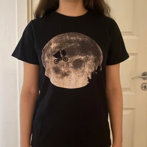 Svart E.T. t-shirt från Dedicated - Svart t-shirt från Dedicated med ikoniskt E.T.-tryck där en cyklist flyger framför en stor måne. Gjord i 100% ekologisk och Fairtrade-certifierad bomull. Klassisk passform och rund hals. Perfekt för dig som gillar film och coola prints.