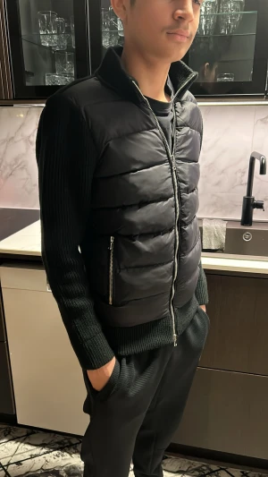 Leon Marcel Cardigen  - Säljer en svart stickad cardigan/tröja med integrerad lätt dunväst framtill. Modellen är både varm och stilren, perfekt för höst, vår eller milda vinterdagar. Cardiganen har: 	•	Mjuk, ribbstickad rygg och ärmar 	•	Blank, vadderad front med två sidofickor 	•	Full längds dragkedja 	•	Bekväm passform som sitter snyggt utan att vara för tight  Helt ny med prislapp, inga defekter, fläckar eller skador. Passar perfekt till vardags, skola, jobb eller en mer dressad streetwear-stil! 