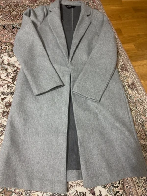 Grå lång kappa från F&F - Snygg grå kappa från F&F med klassisk krage och rak passform. Lång modell med stilrena linjer och två fickor på insidan. Perfekt för dig som gillar minimalistisk och clean stil. Materialet är mjukt och känns som ullblandning.