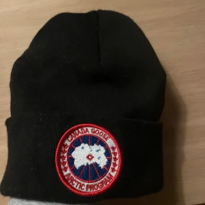 Svart Canada Goose mössa med patch - Svart mössa med klassisk rund patch framtill i rött, vitt och blått. Mössan är ribbstickad och har uppvikt kant. Perfekt för kalla dagar och har en clean, stilren look som funkar till allt.