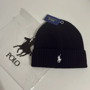 Svart ribbad mössa från Polo Ralph Lauren - Svart ribbstickad mössa från Polo Ralph Lauren med klassisk broderad logga i vitt framtill. Mössan har uppvikt kant och är tillverkad i mjuk ullblandning som håller dig varm. Perfekt accessoar för kyliga dagar med stil.