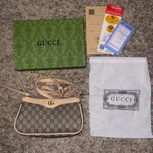 Beige axelväska från Gucci med GG-mönster - Snygg beige axelväska från Gucci med klassiskt GG-mönster i canvas och detaljer i ljust skinn. Väskan har guldfärgad kedja och justerbar axelrem i skinn, samt dragkedja upptill. Perfekt för dig som vill ha en ikonisk och trendig accessoar.