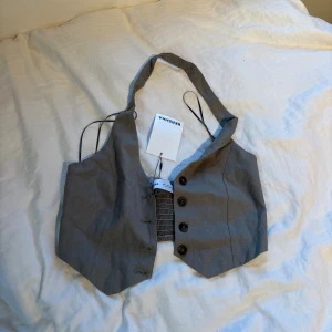 Grå croppad väst från Bershka - Snygg grå croppad väst från Bershka med knappar framtill och elastisk smock baktill. Västen har en v-ringad design och är tillverkad i ett mjukt bomullsmaterial. Grön/grå färg och helt oanvänd!