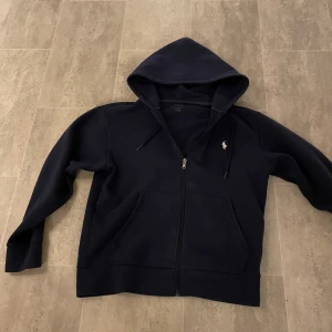 Mörkblå hoodie från Polo Ralph Lauren - Mörkblå hoodie från Polo Ralph Lauren med dragkedja framtill, huva med snörning och klassisk vit logga på bröstet. Funkar helt perfekt .priset går och diskuteras 