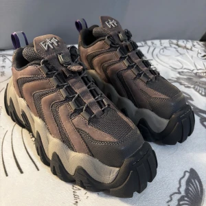 Chunky sneakers från Eytys i brunt - Säljer ett par chunky sneakers från Eytys i mörkbrunt och grått med grov plattformssula. Skorna har mesh och mockadetaljer, snörning och lila dragflik bak. Perfekta för dig som gillar statement-skor med cool design och robust känsla. Obs. Det finns en liten fläck på en skor.