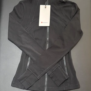 Svart Define jacka från Lululemon storlek 2 - Supersnygg svart Define jacka från Lululemon med figurnära passform och långa ärmar. Jackan har dragkedja framtill, diskreta fickor med dragkedja och coola sömdetaljer som ger en sportig vibe. Materialet är mjukt och stretchigt med nylon och elastan.