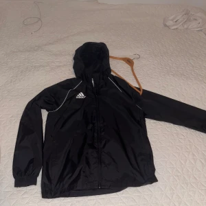 Svart vindjacka från Adidas - Svart vindjacka från Adidas med vit logga på bröstet och vita detaljer längs axlarna. Jackan har huva, dragkedja framtill och långa ärmar med resår vid ärmslut. Perfekt för sportiga dagar eller när du vill ha en chill streetstyle.