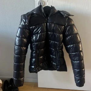 Jacka Nikki modell - Snygg svart pufferjacka från 157 i storlek XXS. Jackan har glansigt material, huva, två dragkedjefickor och markerade sömmar som ger en cool look. Perfekt för kalla dagar och har en modern, lite kortare passform.