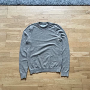 Grå stickad tröja från Strömberg - Snygg grå stickad tröja från Strömberg i storlek S. Tröjan har rund halsringning och ribbade muddar vid ärmslut och nederkant. Perfekt för dig som gillar enkel och stilren design med en neutral färg som passar till allt.