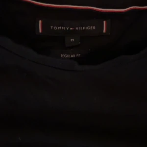 Svart t-shirt från Tommy Hilfiger - Snygg svart t-shirt från Tommy Hilfiger i regular fit. Klassisk rund hals och diskret logga på bröstet. T-shirten har korta ärmar och är gjord i mjuk bomull som känns skön mot huden. Perfekt för dig som gillar stilrena och enkla plagg.