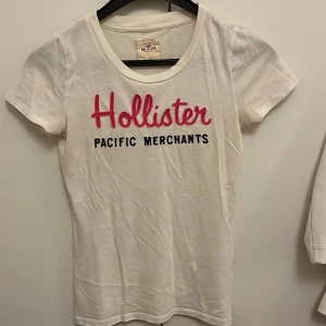 Vit Hollister t-shirt med tryck - Vit t-shirt från Hollister med coolt tryck i rosa och svart på bröstet. Klassisk rund hals och korta ärmar. Perfekt för dig som gillar enkel och stilren design med en touch av färg. Gjord i mjuk bomull som är skön mot huden.