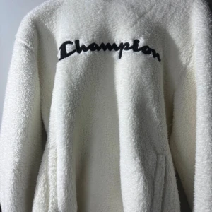Vit fleece-tröja från Champion - Säljer en fluffig och mjuk vit fleece-tröja från Champion med svart broderad logga över bröstet. Tröjan har långärmad design och är perfekt för att hålla värmen under kyliga dagar. Enkel och stilren look som passar till många outfits.