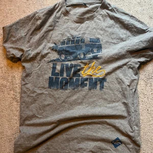 Grå Corona Extra t-shirt med tryck - Säljer en grå t-shirt från Corona Extra i storlek L. Framsidan har ett coolt tryck med en van och texten 'Live the Moment' i blått och gult. Perfekt för dig som gillar Corona 