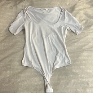 Vit kortärmad body från H&M - En enkel och stilren vit body från H&M med korta ärmar och rundad urringning. Tillverkad i mjuk bomull med knäppning nedtill. Perfekt att styla med jeans eller kjol för en clean look.