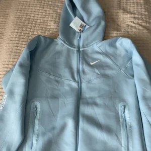 Nike NOCTA ljusblå set - Nike NOCTA set i ljusblå färg med hel dragkedja och huva. Snyggt NOCTA-tryck på ärmen och klassisk Nike Swoosh på bröstet. Hoodien har två fickor med dragkedja och underdelen med dragkedja. Perfekt för chill eller streetwear. Bara att höra av sig vid funderingar😎👊