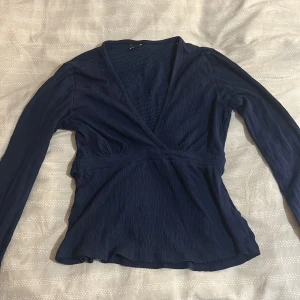 Mörkblå omlottopp med v-ringning från gina tricot - Snygg mörkblå omlottopp med lång ärm och djup v-ringning. Toppen har en lätt strukturerad yta och markerad midja som ger en smickrande siluett. Perfekt att styla med jeans eller kjol för en trendig look.