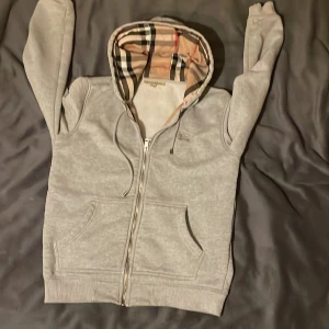 Grå Burberry hoodie med dragkedja - Grå hoodie från Burberry med klassiskt rutigt mönster i huvan och längs insidan. Tröjan har huva med snörning, två fickor fram och hel dragkedja. Perfekt för dig som gillar stilrena detaljer och ikonisk design. Inga defekter eller konstigheter