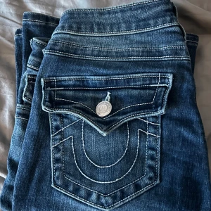 True Religion Joey Low Rise Flare Jeans - Snygga blå jeans från True Religion, modell Joey Low Rise Flare. De har låg midja, utställda ben och klassiska vita kontrastsömmar. Bakfickorna har den ikoniska hästskosömmen och silverfärgade knappar. Perfekt för dig som gillar retrovibbar och coola detaljer. orginalpris 1400kr