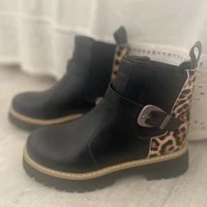 Svarta boots med leoparddetaljer! - Säljer ett par svarta boots med chunky platt sula och rund tå. Baksidan har leopardmönster i bruna och beige toner, samt en snygg metallspänne på sidan. Bootsens ovandel är i syntetmaterial och har dekorativa sömmar runt sulan. Perfekt för dig som gillar att sticka ut.