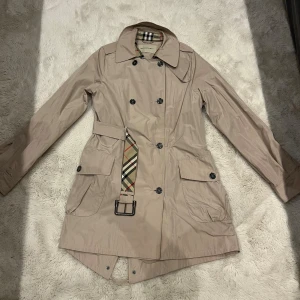 Beige trenchcoat från Burberry - Säljer en beige trenchcoat från Burberry med klassiskt rutigt bälte och foder. Jackan har dubbelknäppning, stora fickor med lock och snygg krage. Materialet är polyester och jackan har långa ärmar. Perfekt för dig som vill ha en ikonisk och stilren look.