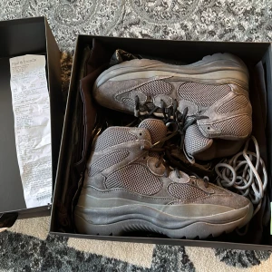 Yeezy Desert Boot Graphite 43 - Säljer ett par Yeezy Desert Boot i färgen graphite. Boots med chunky platt sula, snörning och rund tå. Materialmix av tjock mocka, mesh och nubuck ger en cool och robust vibe. Perfekt för dig som gillar streetwear och vill sticka ut med snygga boots. Ena Original skosnöret är svår o använda men slänger med ett par extra:) vid snabb affär kan jag gå ner i pris:)