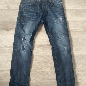Slim fit jeans från Jack & Jones - Mörkblå jeans från Jack & Jones Originals, modell Tim Slim Fit. Byxorna har slitna detaljer och hål på framsidan för en edgy look. Klassisk femficksdesign och raka ben. Perfekt för dig som gillar streetstyle. Skriv för fler frågor med mera
