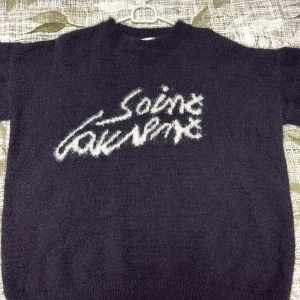 Svart Saint Laurent stickad tröja - Svart stickad tröja från Saint Laurent med vit fuzzy logotyp framtill. Tröjan har rund hals, lång ärm och en loose passform som ger en chill vibe. Materialet är mjukt och fluffigt, perfekt för att hålla dig varm och snygg under kyliga dagar.