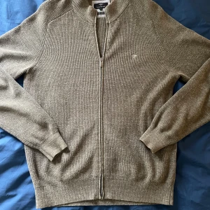 Beige ribbad kofta från Fynch-Hatton - Säljer en beige ribbad kofta från Fynch-Hatton i storlek S. Koftan är tillverkad av superfine bomull, har långärmade ärmar och hel dragkedja framtill. Diskret broderad logga på bröstet och klassisk ståkrage. Perfekt för lager-på-lager under höst och vår.