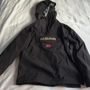 Svart vinterjacka från Napapijri - Svart vinterjacka från Napapijri med stor logotyp på bröstet och norsk flagga. Jackan har huva, halv dragkedja och grå fleecefoder på insidan. Perfekt för kyliga dagar och har en snygg patch på ärmen. Pris kan diskuteras!