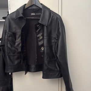 Svart skinnjacka från Zara - Snygg svart skinnjacka från Zara med klassisk krage och raka linjer. Jackan har två stora fickor framtill, knäppning med metallknappar och långärmade ärmar. Storlek S