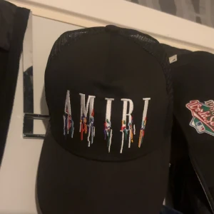 Svart AMIRI keps  - Svart truckerkeps från AMIRI med vit broderad logga framtill och färgglada detaljer som droppar ner från bokstäverna. Perfekt för dig som gillar streetwear och vill sticka ut. Bara att höra av sig vid intresse!😎