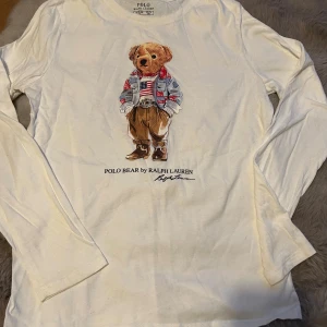 Vit långärmad tröja Polo Ralph Lauren - Vit långärmad tröja från Polo Ralph Lauren med ikoniskt Polo Bear-tryck på bröstet. Tröjan har rund hals och är gjord i mjuk bomull, perfekt för dig som gillar klassisk streetstyle med en twist. Storleken är Xs/160 och passar dig som vill ha en chill look.