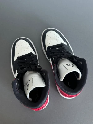 Nike Air Jordan 1 Mid - Nike Air Jordan 1 Mid sneakers med klassisk design i rött, svart och vitt. Skorna har snörning, perforerad tå och rund sula. Swoosh-logga i rött på sidan och Air Jordan Wings på ankeln. Tillverkade i skinn med svart foder och vit platt sula.