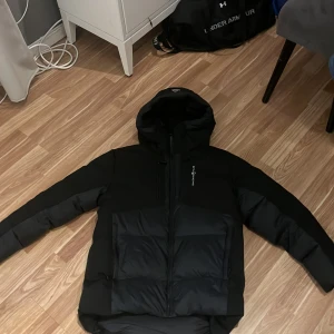 Svart Sail Racing dunjacka med huva - Svart dunjacka från Sail Racing med huva och dragkedja. Jackan har quiltad design, lång ärm och är fylld med Allied Feather + Down (500 FP). Snyggt märke på ärmen och stilren look som håller dig varm under kalla dagar. Aldrig andvänd/ nyskick. Storlek=170cm