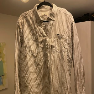 Skjorta Sons of Owen - Snygg randig skjorta från Sons of Owen i storlek XL. Skjortan är beige med smala grå ränder, har två bröstfickor.
