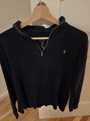 Marinblå half zip från Polo Ralph Lauren - Snygg marinblå långärmad tröja från Polo Ralph Lauren med klassisk broderad logga på bröstet. Tröjan har half zip-dragkedja och hög krage, perfekt för lager-på-lager. Tillverkad i mjuk bomull för en skön känsla.