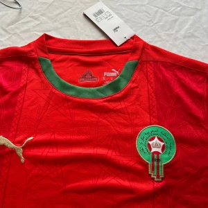 Marocko fotbollströja Puma röd/grön - Säljer en officiell Marocko fotbollströja från Puma i rött med gröna detaljer och guldiga Puma-loggor. Tröjan har korta ärmar, rund hals och är tillverkad i lätt, ventilerande polyester med subtila stjärnmönster. Landslagsemblemet är broderat på bröstet.