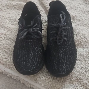 Adidas svarta sneakers - Svarta Adidas  sneakers med stickad ovandel och mönstrad textur. Skorna har rund tå, platt sula och snörning med tjocka svarta och grå skosnören. Diskret röd detalj på hälen och ribbad sula ger en cool look. Perfekt för dig som gillar streetstyle.