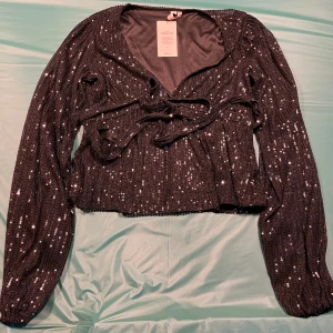 Svart glittrig omlottopp från Nelly - Säljer en svart omlottopp från Nelly med glittriga paljetter över hela tyget. Helt oanvänd med lappen kvar!