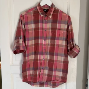 Rutig röd skjorta från Dr Denim - Snygg rutig skjorta från Dr Denim i rött, rosa och beige med inslag av grått. Skjortan har klassisk krage, bröstficka och knappar framtill. Ärmarna kan rullas upp och fästas med knapp. Perfekt för dig som gillar avslappnad stil med lite färg.
