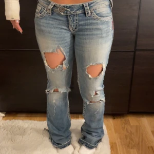 Silver Jeans ljusblå bootcut jeans - Säljer ett par ljusblå bootcut jeans från Silver Jeans med slitna detaljer och hål framtill. Jeansen har broderade bakfickor med vit tråd och klassisk femficksdesign. Materialet är jeans med snygga kontrastsömmar och låg midja. Dom är super sköna och inte stela, storleken är ungefärligde är köpta i Thailand🥰midjemått 39cm innerbenslängden är ca 84, de passar upp till 174 skulle jag säga. De är ganska slitna baktill på hälen också kan därför tänka mig att sänka priset en del! Ge gärna prisförslag