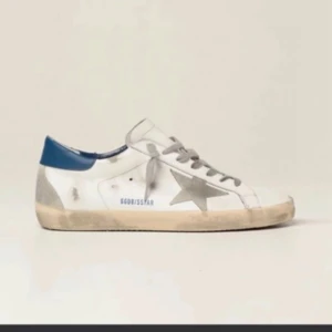 Golden Goose vita sneakers med blå detalj - Golden Goose sneakers i vitt skinn med grå stjärndetalj på sidan, grå snörning och blå hälkappa. Sulan är beige och har en vintage-look. Perfekta för dig som gillar en avslappnad och trendig stil. 46