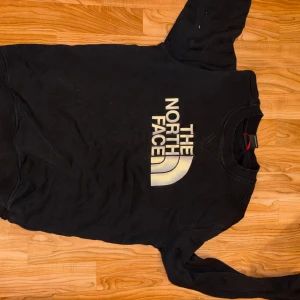 Svart sweatshirt från The North Face - Svart sweatshirt från The North Face med stort vitt logotryck på bröstet och mindre logga på ryggen vid axeln. Tröjan har rund hals, långa ärmar och är gjord i mjuk bomull. Perfekt för chill dagar eller när du vill ha en clean streetstyle.